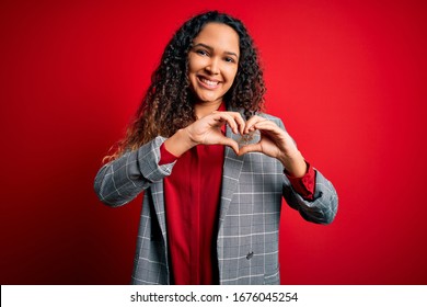 Young beautiful businesswoman with curly hair wearing elegant jacket over red background smiling in love showing heart symbol and shape with hands. Romantic concept.