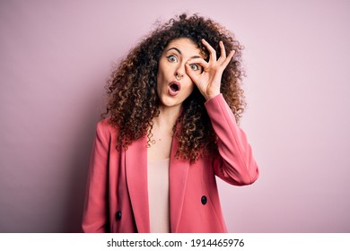 Young beautiful businesswoman with curly hair and piercing wearing elegant jacket doing ok gesture shocked with surprised face, eye looking through fingers. Unbelieving expression.