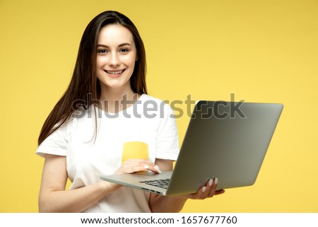 Young beautiful businesswoman with dark short hair in white shirt happily working on laptop over yellow background