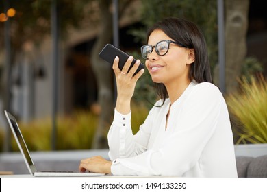 Young Beautiful Businesswoman In Eyeglasses Smiling And Recording Audio Message On Mobile Phone While Sitting At The Table With Laptop Outdoors