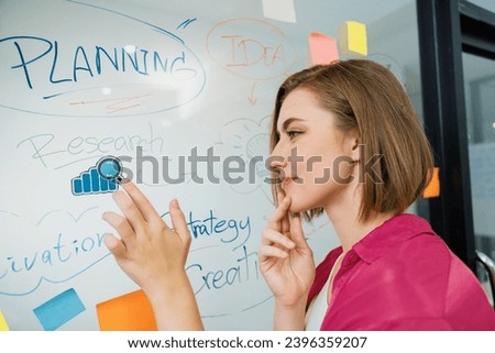 Young beautiful businesswoman putting sticker on glass board while finding a solution to solve financial problems by using mind map and colorful sticky note. Creative business concept. Immaculate.