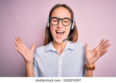 Young beautiful call center agent woman wearing glasses working using headset celebrating mad and crazy for success with arms raised and closed eyes screaming excited. Winner concept