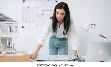 Young beautiful caucasian architect puts blueprint on the table and compare with house model while inspect hos model construction carefully at modern office with blueprint striped behind. Immaculate. - Powered by Shutterstock