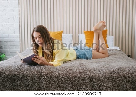 A young beautiful caucasian blonde woman in a yellow shirt is lying in bed and reading a book. Self-isolation. Lock down. Introvert