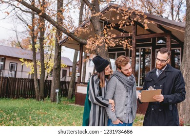 Young beautiful caucasian couple, stylish, dressed in worm outdoor apparel talking to good-looking male propery owner, businessman decides to sell a house, property investments