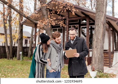 Young beautiful caucasian couple, stylish, dressed in worm outdoor apparel talking to good-looking male propery owner, businessman decides to sell a house, property investments