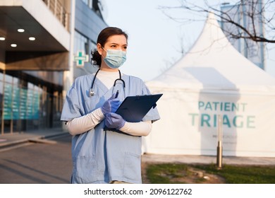 Young beautiful caucasian female NHS doctor in front of emergency ambulance clinic,next to white patient admission triage tent for receiving infected Coronavirus patients,COVID-19 quarantine hospital