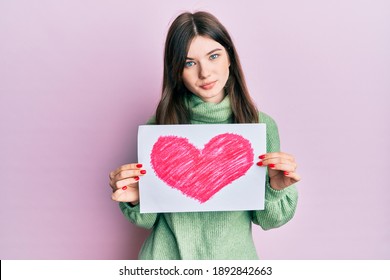 Young beautiful caucasian girl holding heart draw relaxed with serious expression on face. simple and natural looking at the camera. 