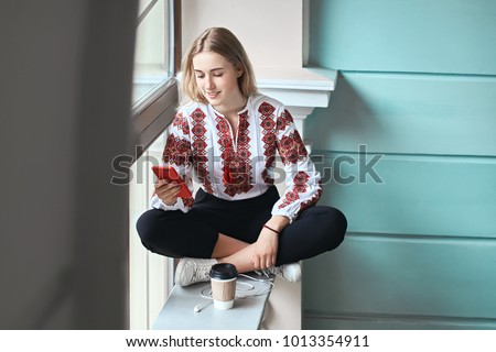 Young beautiful caucasian girl student wearing a vyshyvanka, a traditional Ukrainian embroidered shirt checks her smartphone while drinking coffee between lectures