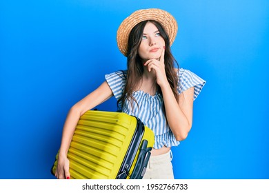 Young beautiful caucasian girl wearing summer dress and holding cabin bag serious face thinking about question with hand on chin, thoughtful about confusing idea 