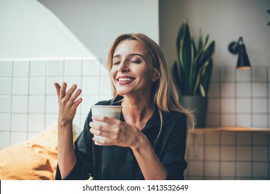 Young beautiful Caucasian happy smiling girl holding cup of hot coffee and smelling it during break in cafeteria, positive female enjoying aroma sniff of caffeine beverage feeling good in cafe