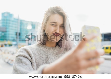 Young beautiful caucasian purple grey hair woman outdoor in the city outdoor in city back light taking selfie with smart phone hand hold doing grimace - vanity, social network, sharing concept