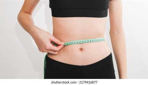 Young Beautiful Caucasian Teen Girl In A Fitness Suit Measures Her Waist With A Measuring Tape With One Hand On The Background Of A White Wall, Side View. Concept Of Sports, Fitness And Modern Lifesty