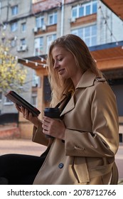 Young Beautiful Caucasian Woman In Beige Stylish Coat Using Tablet Outdoors On Sunny Day, Reading Ebook. Attractive Female Street Portrait At Fall Or Spring. Autumn Lifestyle, Modern Technology