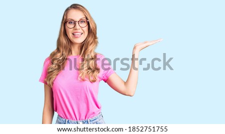 Young beautiful caucasian woman with blond hair wearing casual clothes and glasses smiling cheerful presenting and pointing with palm of hand looking at the camera. 