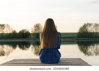 young beautiful caucasian woman in blue dress sits at wooden pier on river bank in summer at sunset, green trees and blue sky in background, melancholy concept, traveling along in vacation