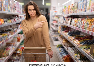 young beautiful caucasian woman with a disgruntled look in the supermarket, the index finger of the hand is directed to the shelf with the goods. Copy space. - Shutterstock ID 1945539976