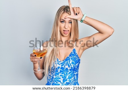 Young beautiful caucasian woman drinking a glass of white wine making fun of people with fingers on forehead doing loser gesture mocking and insulting. 