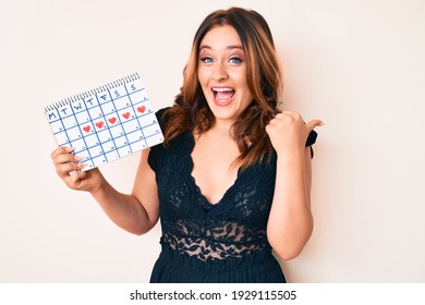 Young beautiful caucasian woman holding heart calendar pointing thumb up to the side smiling happy with open mouth 