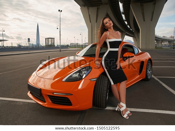 長いまっすぐな髪を持ち オレンジ色のスポーツカーの近くでポーズをとっている若い美しい白人女性 屋外ファッションのポートレート の写真素材 今すぐ編集