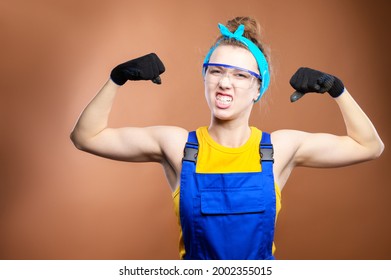 Young Beautiful Caucasian Woman Repairman Worker With Blue Eyes In Uniform Showing Arm Muscles, Proudly Smiling And Making A Strong Grimace. Contractors And Wise Construction Superintendents.