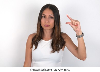 Young Beautiful Caucasian Woman Wearing White Top Over White Background Purses Lip And Gestures With Hand, Shows Something Very Little.