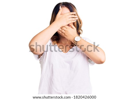 Young beautiful caucasian woman wearing casual white tshirt covering eyes and mouth with hands, surprised and shocked. hiding emotion 