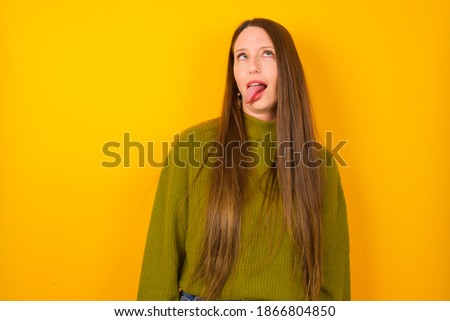 Young beautiful Caucasian woman wearing knitted sweater against yellow background  showing grimace face crossing eyes and showing tongue. Being funny and crazy