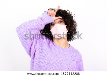young beautiful caucasian woman wearing pink medical mask standing against white wall, making fun of people with fingers on forehead doing loser gesture mocking and insulting.