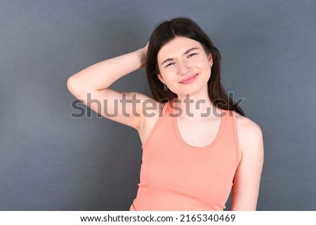 young beautiful Caucasian woman wearing orange shirt over studio grey wall being confused and wonders about something. Holding hand on head, uncertain with doubt. Pensive concept.