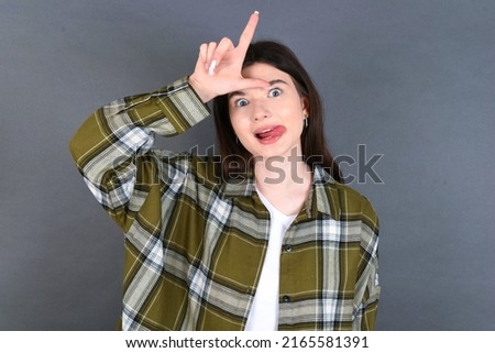 young beautiful Caucasian woman wearing overshirt over studio grey wall, gestures with finger on forehead makes loser gesture makes fun of people shows tongue