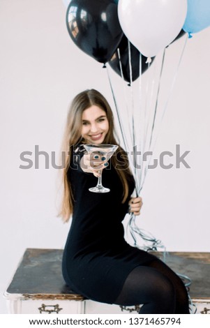 Young beautiful celebrating woman in black dress posing with cocktail in hand and balloons, sitting on the wooden table. Beautiful model portrait . Bright make up. Attractive girl smiling