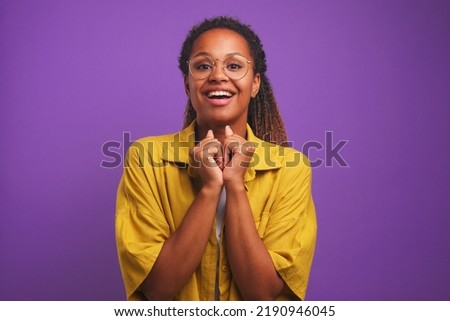 Young beautiful cheerful African American woman smiles excitedly and looks at camera clenching fists raises hands to chin in anticipation of long-awaited meeting stands on studio purple background