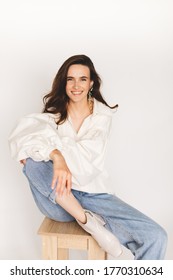 Young beautiful cheerful girl sitting on chair wearing white blouses and jeans on the white background. Young woman smiling very happy surprised holding leg being amazed. Girl hold head and smiling.