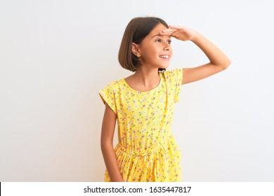 Young beautiful child girl wearing yellow floral dress standing over isolated white background very happy and smiling looking far away with hand over head. Searching concept.