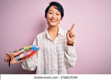 Young Beautiful Chinese Artist Woman Painting Using Paintbrush And Palette With Colors Surprised With An Idea Or Question Pointing Finger With Happy Face, Number One