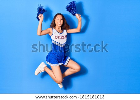 Young beautiful chinese girl smiling happy wearing cheerleader uniform. Jumping with smile on face using pompom over isolated blue background
