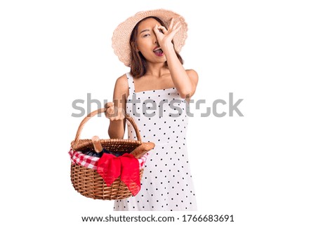 Young beautiful chinese girl wearing summer hat holding picnic basket smiling happy doing ok sign with hand on eye looking through fingers 