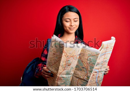 Young beautiful chinese tourist woman holding city map over isolated red background with a happy face standing and smiling with a confident smile showing teeth