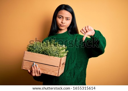 Young beautiful chinese woman holding garden box plants over isolated yellow background with angry face, negative sign showing dislike with thumbs down, rejection concept