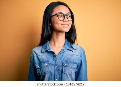 Young Beautiful Chinese Woman Wearing Casual Denim Shirt Over Isolated Yellow Background Looking Away To Side With Smile On Face, Natural Expression. Laughing Confident.