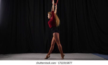 Young Beautiful Circus Actress, A Curly Aerial Gymnast, Performs Elements On An Air Ring Against Black Backstage. Performance On Stage, Theater. Beautiful Blonde Girl Is Engaged In Aerial Hoop.