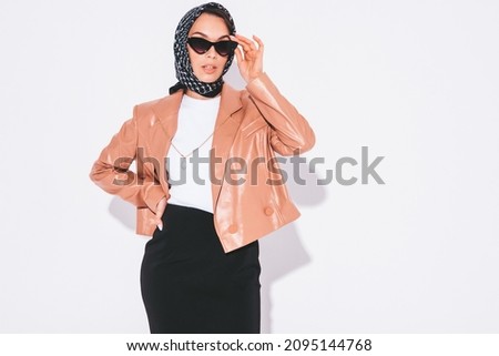 Young beautiful confident brunette female in nice trendy clothes.Sexy carefree woman posing near white wall in studio with head scarf. Fashionable model with bright evening makeup 