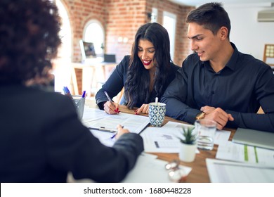 Young Beautiful Couple Applying For Mortgage. Sitting Smiling Happy Meeting With Real State Agent Signing Mortgage Loan At Bank