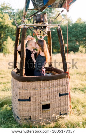 Young beautiful couple in black clothes, kissing in the basket of hot air balloon, enjoying their first fly in warm summer sunrise in the field