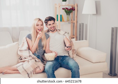 Young beautiful couple of blondie and brunette guy are watching movie under the beige plaid, eating popcorn at light cozy home