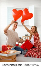 Young beautiful couple celebrates Valentine 's Day at home ,comfortably sitting with blanket , pizza and glasses of wine . They hold red heart-shaped balloons and gifts. Love and happiness. Lovers. 