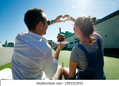 Young Beautiful Couple Eating Fast Food By The Pool, Loving Guy And Girl Eating Burger And Smiling At Each Other