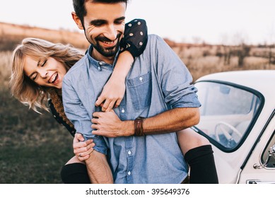 Young Beautiful Couple Hugging Each Other, Sitting On A Small White Car In Beautiful Evening Light. Stylish Guy With A Beard And Blond Girl Laughing