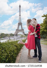 Young Beautiful Couple Kisses Covering Their Faces With A Straw Hat In Front Of Eiffel Tower In Paris. A Girl In A Red Dress With White Polka Dots And A Guy In A Red Plaid Shirt. Romantic Summer.